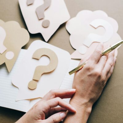 Photo of paper question marks with hands and a pen suggesting curiosity and inquiry.